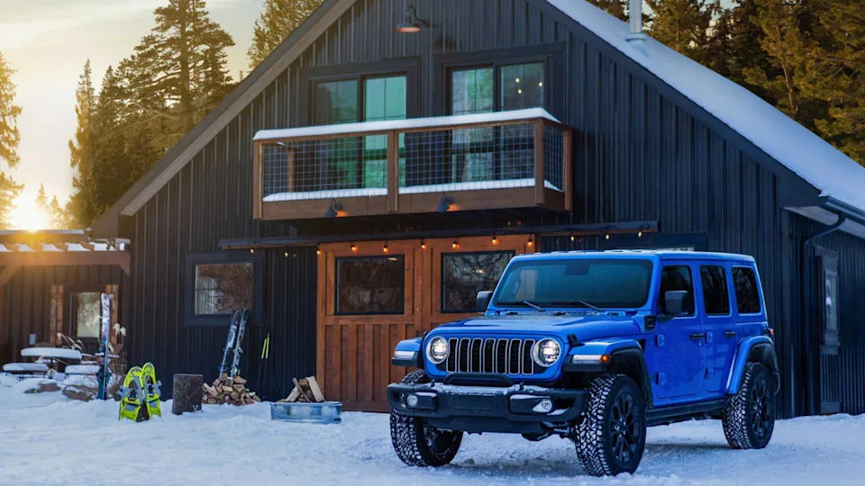 Static front 3/4 shot of a blue 2025 Jeep Wrangler 4xe parked outside of a cabin in the snow.