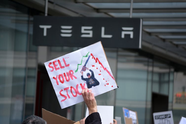 protester outside a tesla branch holds up a sign reading 'sell your stock'
