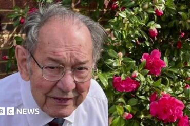 An elderly man with mid-length grey hair. He is stood in front of a bunch of pink flowers wearing a white shirt and tie.