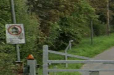Google A large silver gate blocks the entrance to the bridleway, which is long and flanked by trees and bushes.