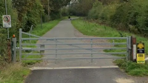 Google A large silver gate blocks the entrance to the bridleway, which is long and flanked by trees and bushes.