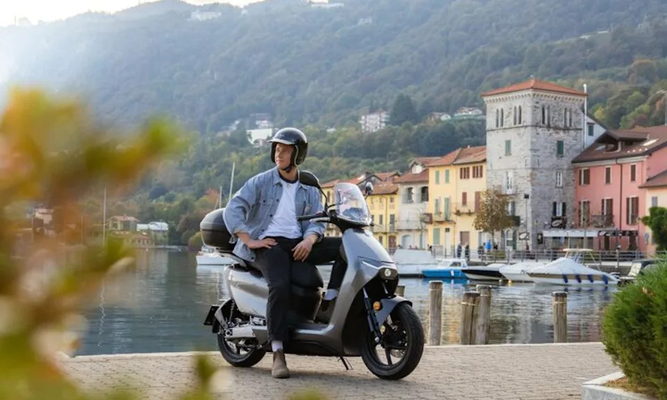 A single rider on a Zero LS1 scooter stopped n a stone pathway looking backward with sonte buildings, yacht dockage, and mountains in the background.