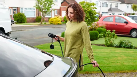 Getty Images Woman wearing a green jumper is taking her charger to her car. She has curly brown hair. 