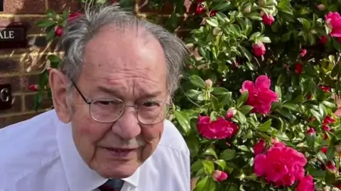 Family Handout An elderly man with mid-length grey hair. He is stood in front of a bunch of pink flowers wearing a white shirt and tie.
