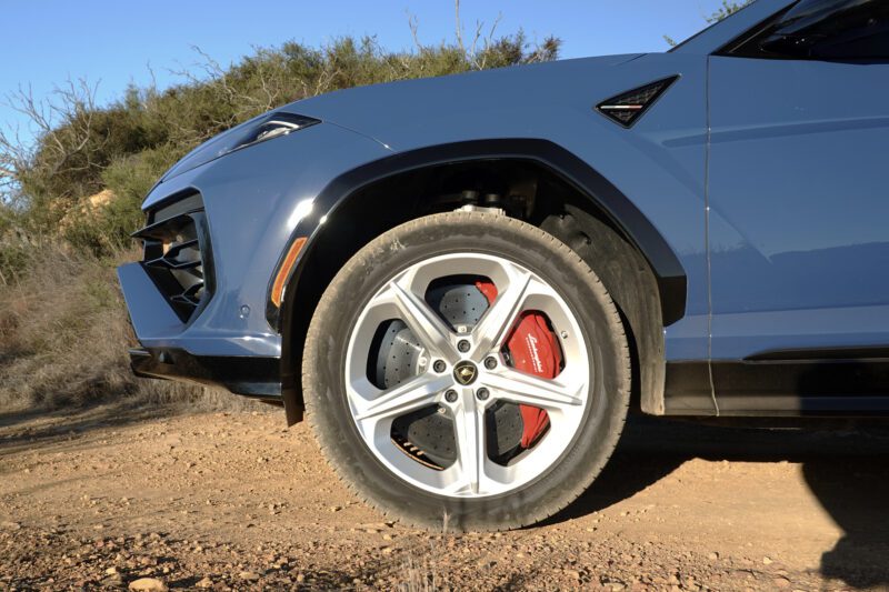 Close-up of the front left wheel of a blue SUV with red brake calipers, parked on a dirt path with vegetation in the background—perfect for capturing details highlighted in the 2025 Lamborghini Urus SE Review: Heavy Hitter Delivers Plug-In Hybrid Punch.