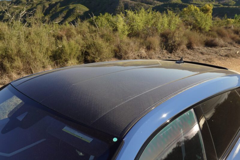 Close-up view of a car roof with a carbon fiber finish, parked outdoors near shrubs and hills under daylight—a style detail seen in the 2025 Lamborghini Urus SE Review: Heavy Hitter Delivers Plug-In Hybrid Punch.