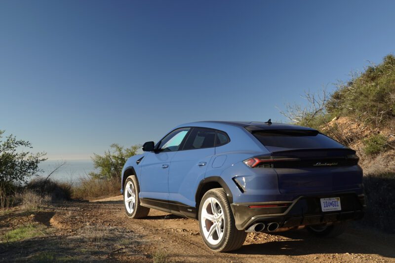 A blue 2025 Lamborghini Urus SE, the heavy hitter that delivers a plug-in hybrid punch, sits on a dirt road with striking white wheels, parked beside a hillside under a clear blue sky.