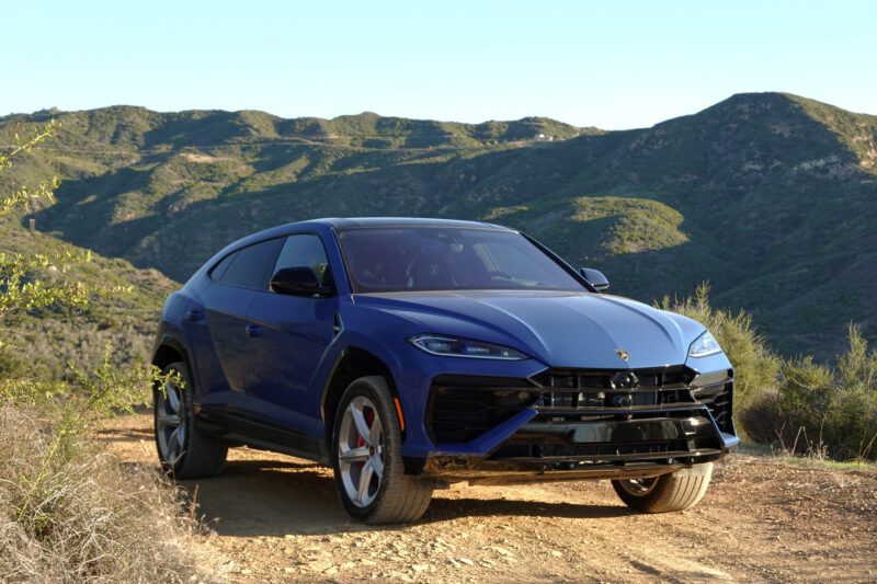 A blue 2025 Lamborghini Urus SE is parked on a dirt path with green hills and blue skies in the background, showcasing this heavy hitter’s plug-in hybrid punch.