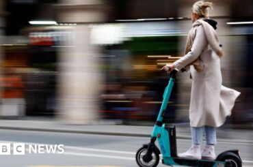 Getty Images A woman riding on an e-scooter on the road. She is wearing a long cream-coloured coat and scarf, blue jeans and white high-top trainers. She has blonde hair clipped back with a clasp.
