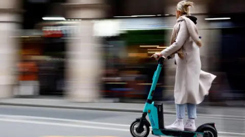 Getty Images A woman riding on an e-scooter on the road. She is wearing a long cream-coloured coat and scarf, blue jeans and white high-top trainers. She has blonde hair clipped back with a clasp.