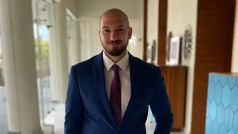 Youssef Kamal Youssef Kamal with a beard and shaved head wears a blue suit and tie