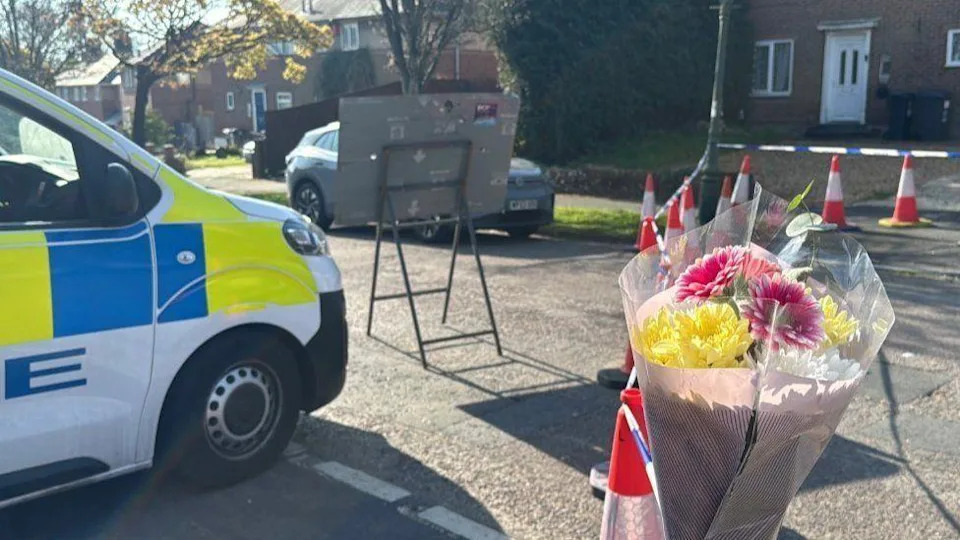 Pink, yellow and white flowers attached to police tape on a traffic cone in a road with a police van to the left and homes in the distance - it is a sunny day.