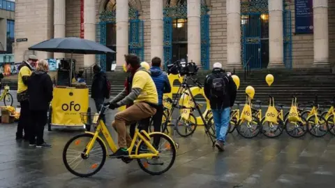 Ofo A number of bright yellow bikes are lined up outside Sheffield City Hall. There is a man on one of the bikes wearing a matching yellow t-shirt and brown trousers cycling past