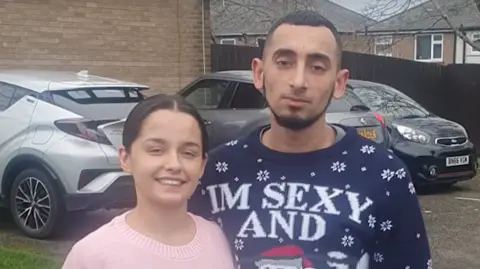 WMFS A woman in a pink top and wearing a pink top, with tied-back dark hair; and a man with short dark hair and beard, wearing a blue Christmas jumper, stand in front of some cars parked by a house. They are both looking at the camera.