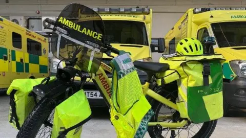 Acute Ambulance & Medical Services Ltd A bright yellow emergency response bicycle equipped for ambulance duties. It has large pannier bags on both the front and rear sides, also in fluorescent yellow with reflective strips for safety. The front panniers are mounted on either side of the wheel, and the rear panniers appear to carry medical supplies. There are ambulances in the background.