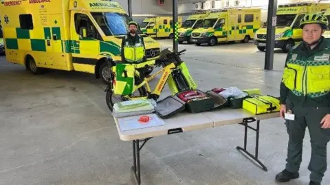 Acute Ambulance & Medical Services Ltd An ambulance station with several yellow and green emergency ambulances parked in the background. In the foreground, there is a table displaying various medical and emergency response equipment, including bags and folded items. Next to the table is a bright yellow ambulance bicycle equipped with panniers and marked with the word “AMBULANCE”. 
