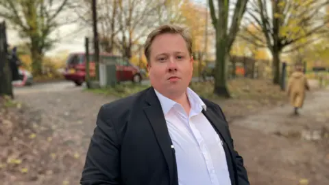 BBC Alistair Brown pictured in Florence Park in Oxford - he is wearing a white shirt and black jacket and looking at the camera with a serious expression.