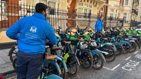 Two men in blue anoraks are moving bikes out of the carriage way onto the pavement.