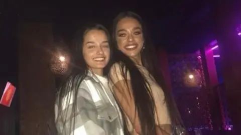 Handout Two women smiling at the camera, leaning into each other, inside a bar.