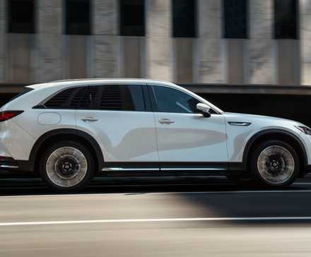 Dynamic side profile shot of a white 2025 Mazda CX90 driving in a city.