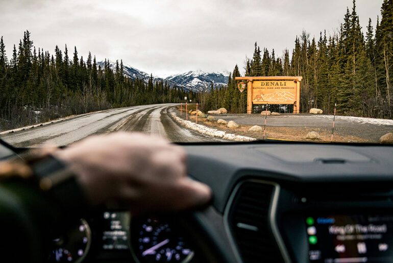The view looking down a highway in Alaska through the windshield of a personal vehicle with a road sign for Denali park sits in the distance.