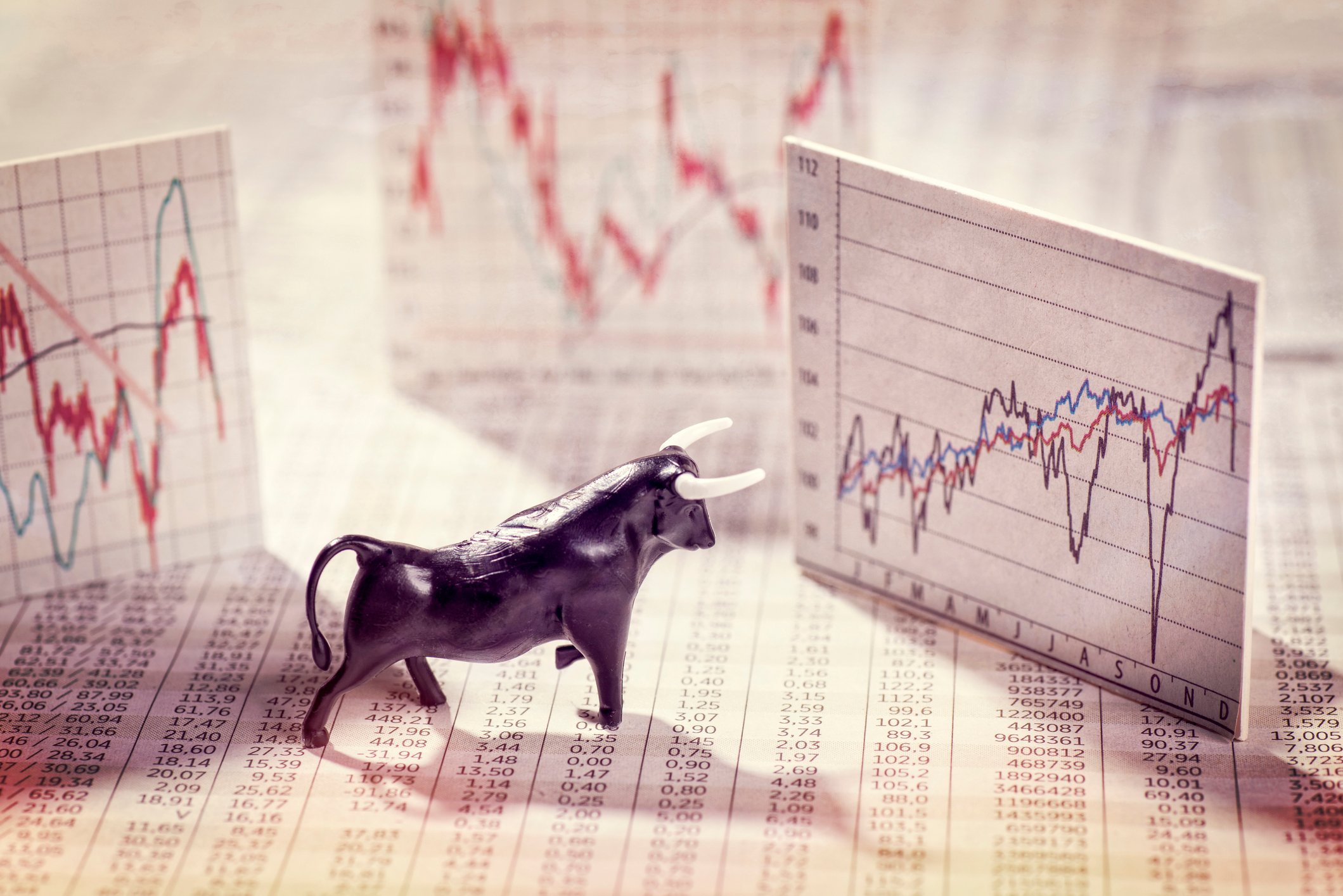A bull figurine stands on newsprint and looks at index cards showing stock price charts.