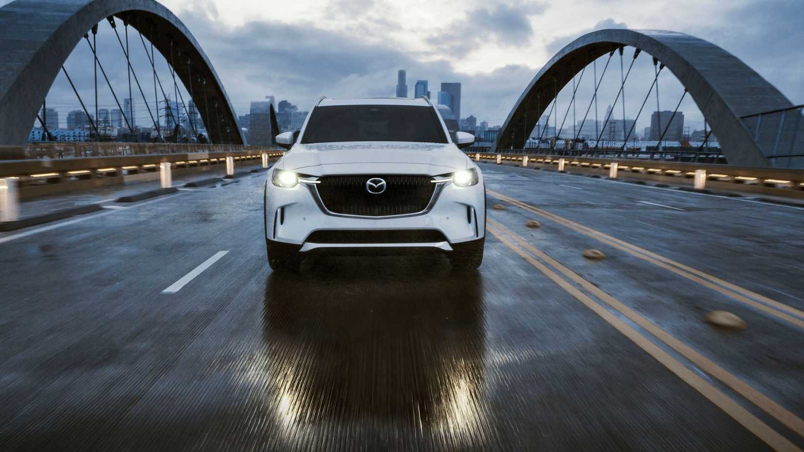 Dynamic front-end shot of a white 2026 Mazda CX-90 driving over a bridge with a cityscape in the background.