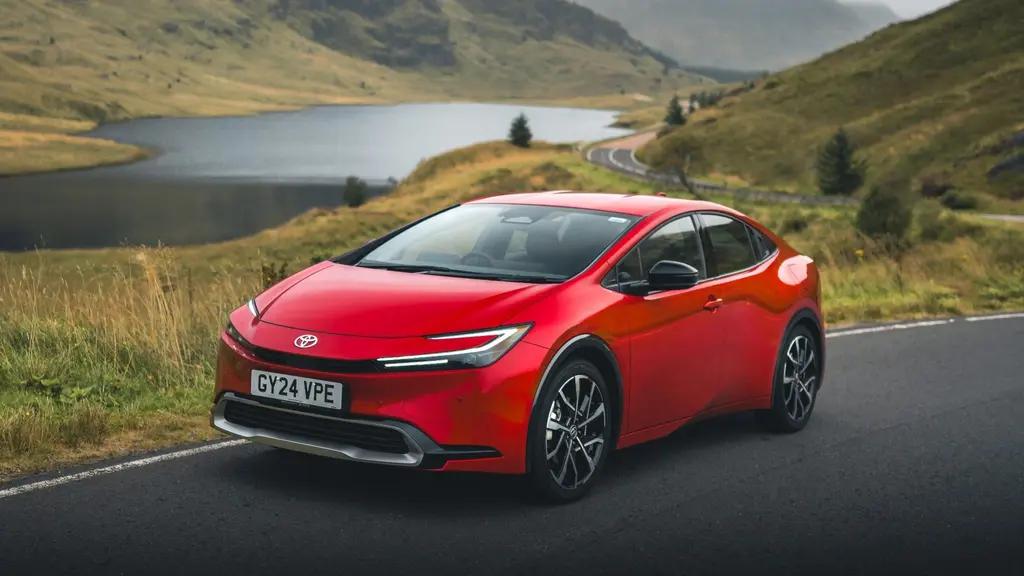 A red Toyota Prius parked beside a lake in a hilly UK scene.