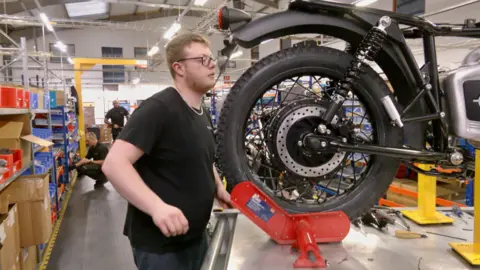 A man with glasses and brown hair is near the back of a motorbike, which is on a raised surface.