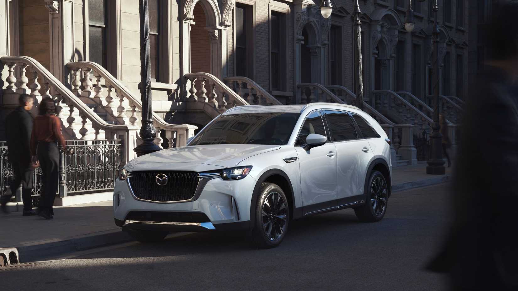 Static front 3/4 shot of a white 2026 Mazda CX-90 parked on a street outside some houses.