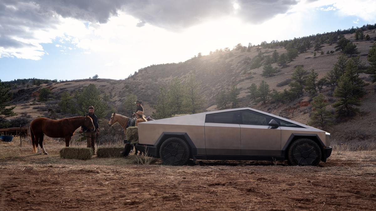 A tan Tesla Cybertruck parked in a rustic, rural setting next to two horses and hay bales, with hills in the background.
