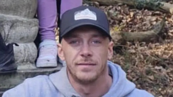 Joey Johnstone poses for a photograph outdoors. He wears a baseball cap and a light grey hoodie and has a short beard.