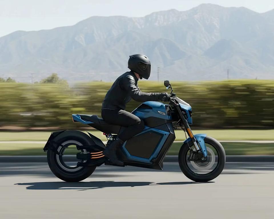Person wearing black helmet and gear riding a blue electric motorcycle on a road with mountains in the background.