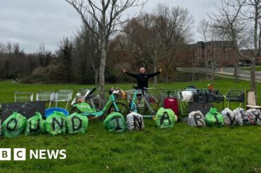 Mark Rowlands In a green park, Mark Rowlands poses with his arms out as he stands behind a row of rubbish backs with the letters