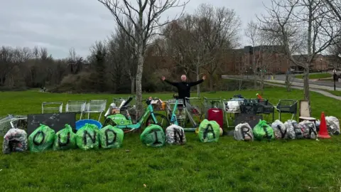 Mark Rowlands In a green park, Mark Rowlands poses with his arms out as he stands behind a row of rubbish backs with the letters 'FOUND IN A RIVER' written on them, as well as a row of shopping trolleys, e-bikes and e-scooters. 