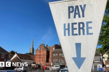 BBC A white sign at a car park in Salisbury, Wiltshire, which reads "pay here" in blue writing, with a blue arrow pointing down. The sign is shaped like an upside down triangle. Cars can be seen parked in the city car park behind, which is surrounded by older buildings and a large spire can be seen in the background. It is a sunny day.