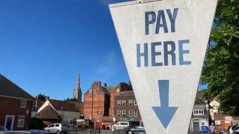 BBC A white sign at a car park in Salisbury, Wiltshire, which reads "pay here" in blue writing, with a blue arrow pointing down. The sign is shaped like an upside down triangle. Cars can be seen parked in the city car park behind, which is surrounded by older buildings and a large spire can be seen in the background. It is a sunny day.