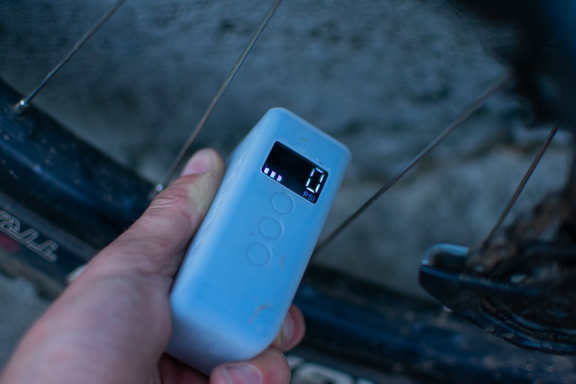 A hand holding a small digital tire pressure gauge displaying a reading of 0 PSI, with a bicycle wheel visible in the background.