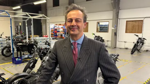 A man wearing a tie, blue shirt and pinstripe jacket is inside, looking at the camera. Motorbikes are behind him.