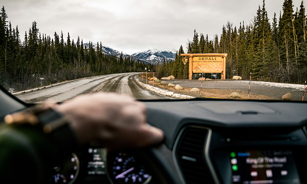 The view looking down a highway in Alaska through the windshield of a personal vehicle with a road sign for Denali park sits in the distance.