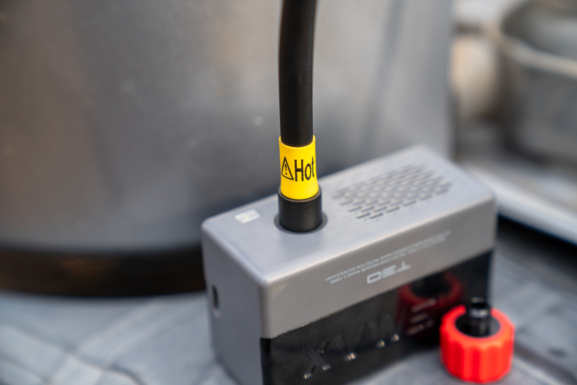 A close-up of a device with a black hose labeled "Hot" on a gray base, alongside a red cap, with a blurred background suggesting an outdoor or workshop setting.