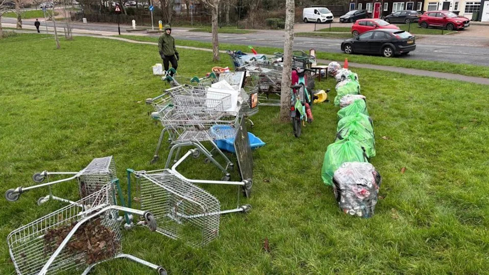 A row of shopping trolleys and rubbish bags sit on a patch of grass next to a road