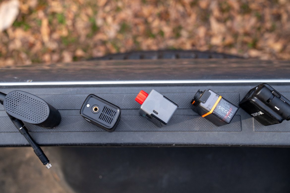 A close-up view of various small electronic devices arranged on a textured surface, including a speaker, a camera module, and two battery packs, with a background of fallen leaves.