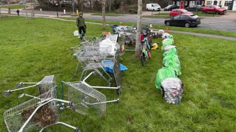 Mark Rowlands A row of shopping trolleys and rubbish bags sit on a patch of grass next to a road