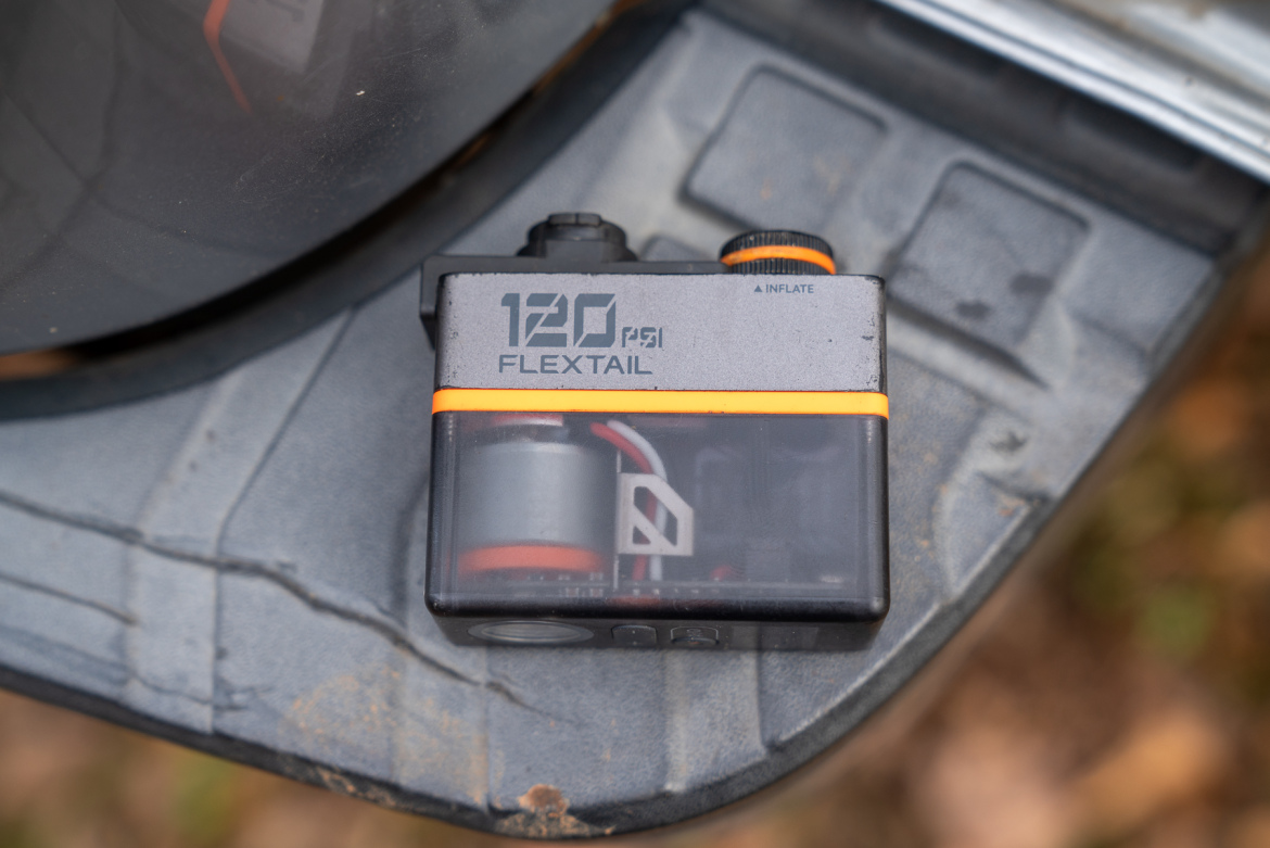 A compact black and orange air pump labeled "120 PSI FLEXTAIL" positioned on a textured surface, with visible mechanical components inside a transparent casing.