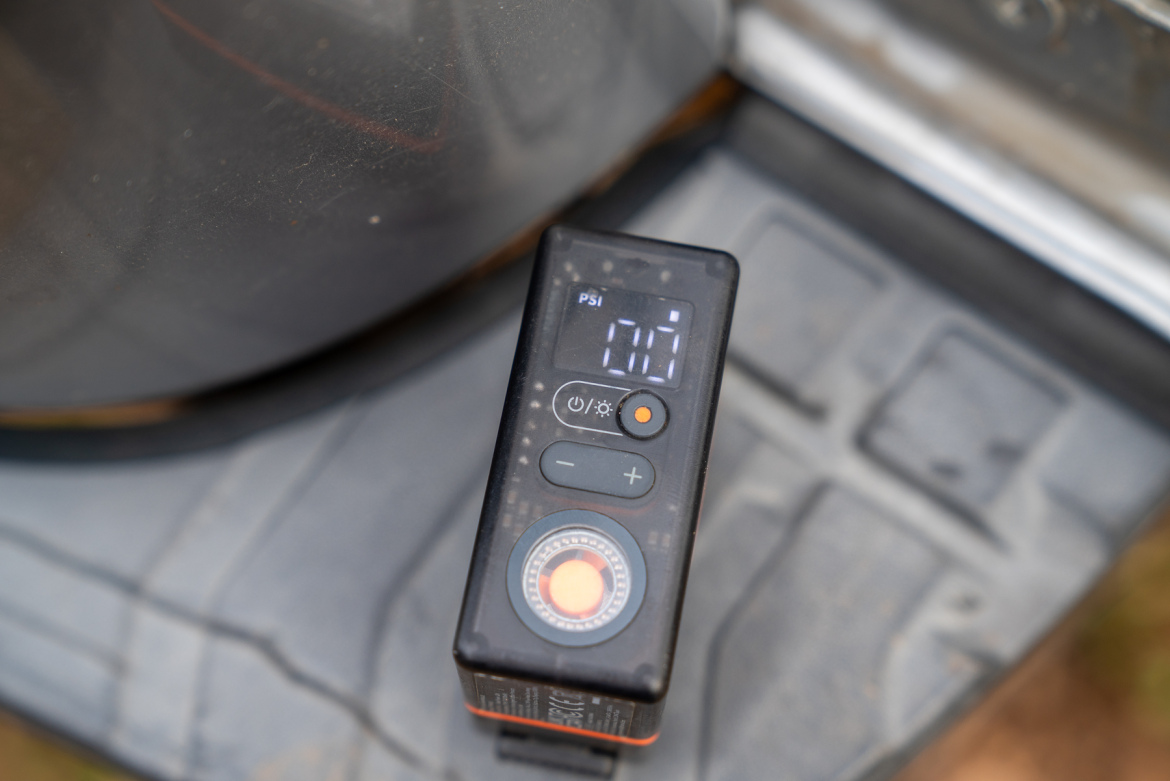A digital air pressure gauge resting on a textured surface, displaying a reading of 00 PSI. The device features a circular pressure adjustment dial and control buttons. In the background, part of a vehicle tire is partially visible.