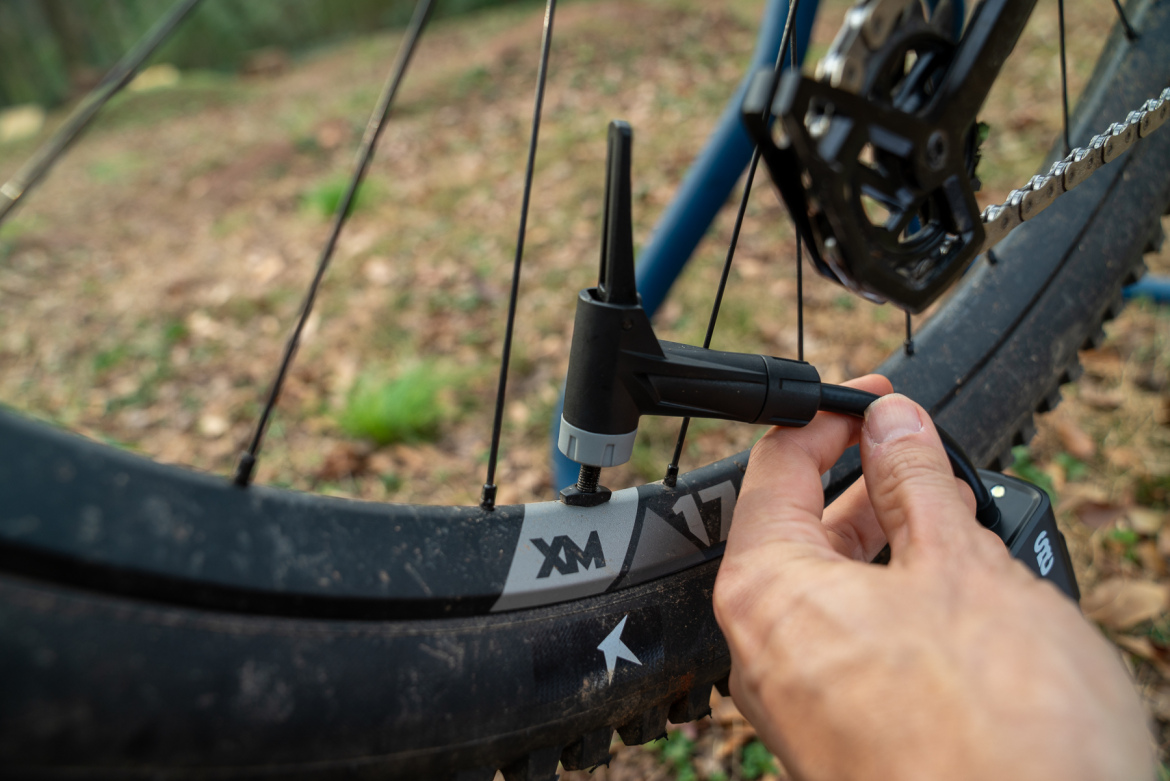 A close-up image of a hand using a tool to adjust the air pressure in a mountain bike tire, set against a natural outdoor background with leaves and grass.