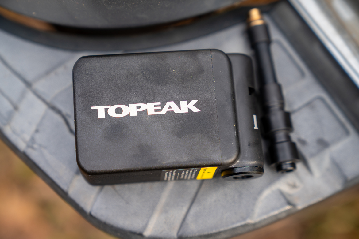 Black Topeak pump resting on a textured surface, with a visible hose attachment next to it. The top of the pump features the "TOPEAK" logo in white letters.