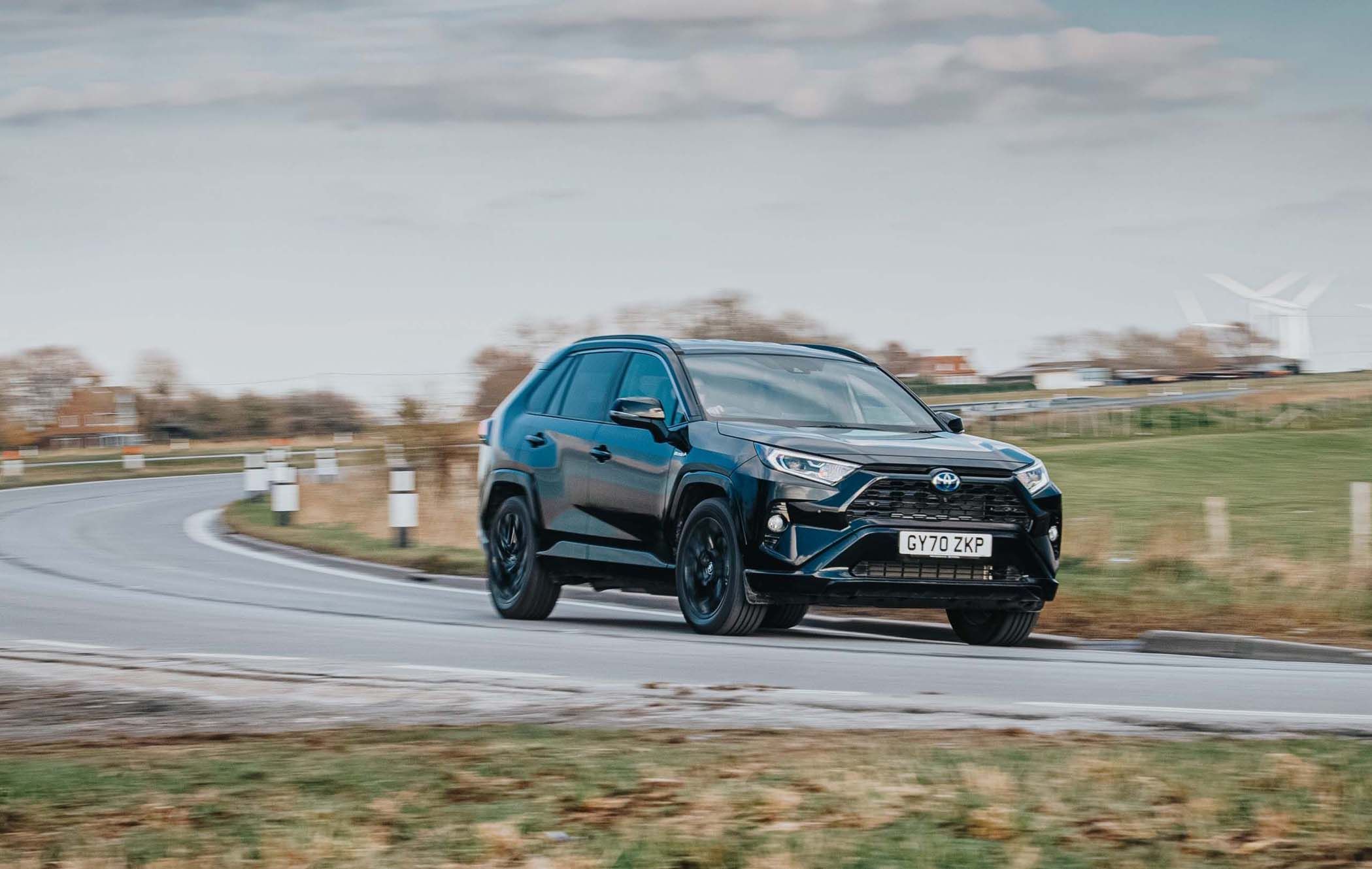 A black Toyota RAV4 driving on a curved rural road, surrounded by grass fields and cloudy skies.