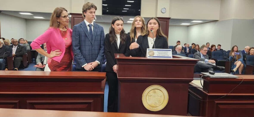 Five people stand in front of a large group of people and speak behind a podium with a mic. 